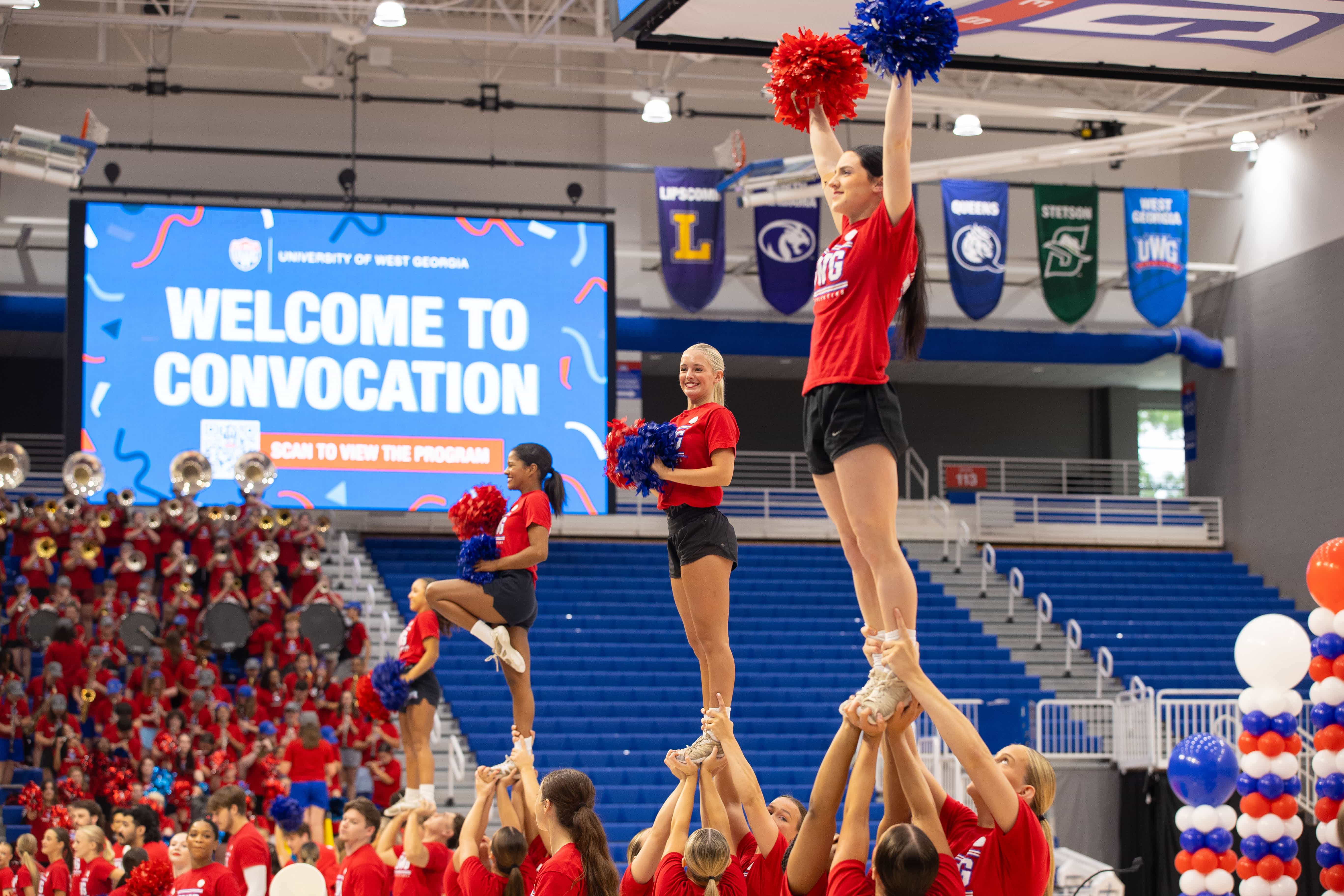 UWG cheer team in red on the basketball court with the band also in red playing music in the bleachers.