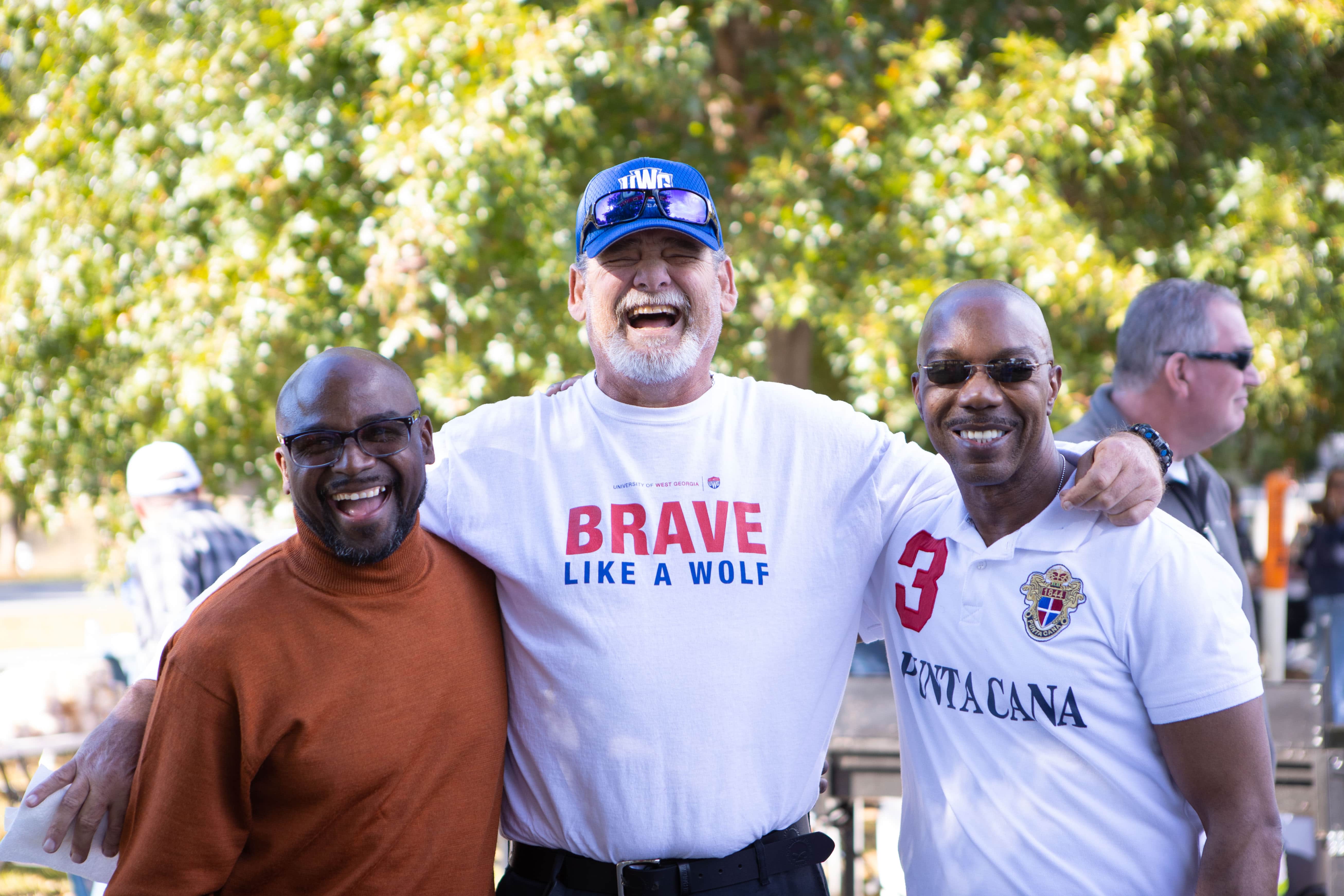 Three UWG alumns standing together for a photo. The center person is wearing a white shirt with "Brave Like a Wolf" written on the front, and a UWG hat.