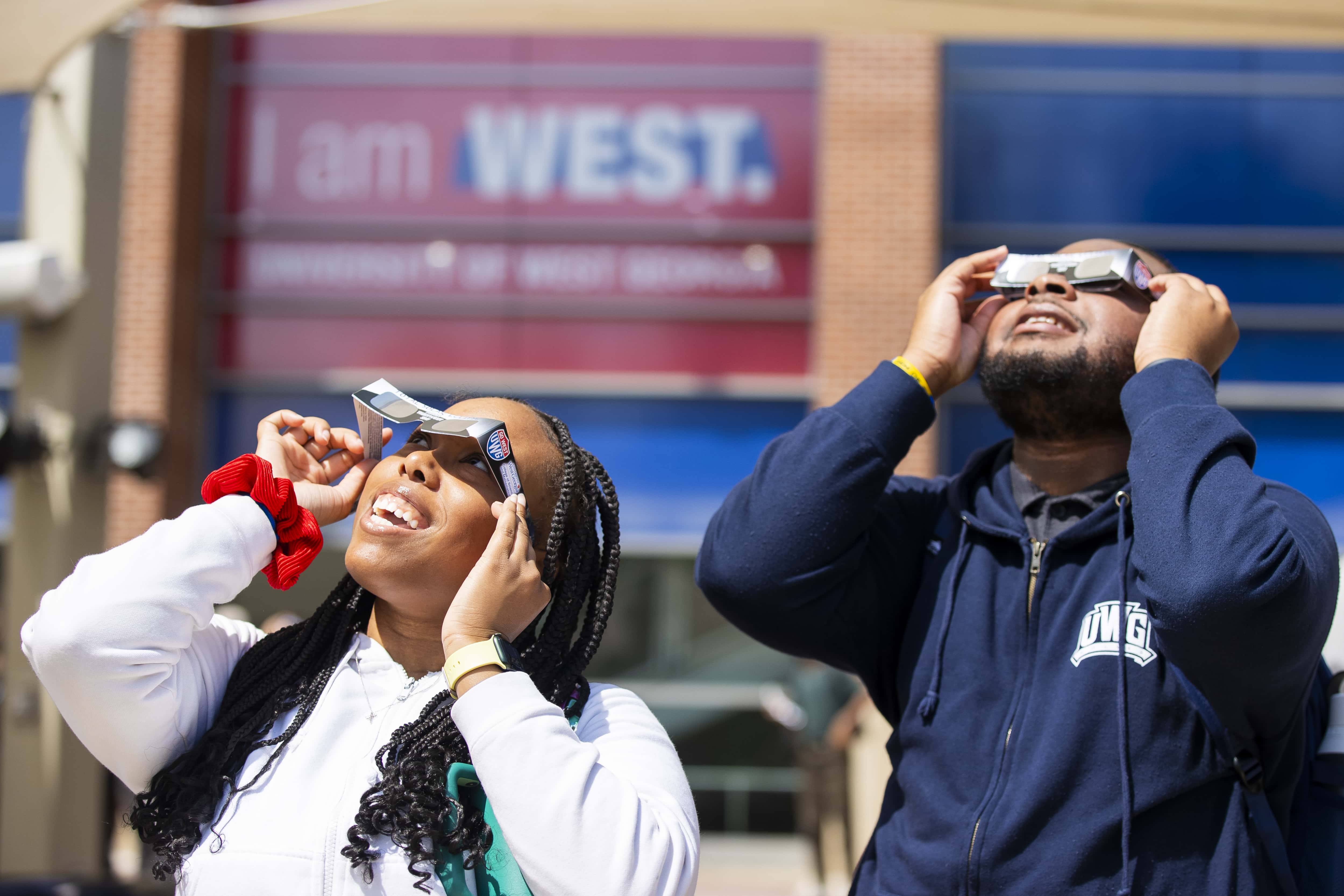 Students standing outside the Campus Center looking at the solar eclipse with UWG branded eclipse glasses.